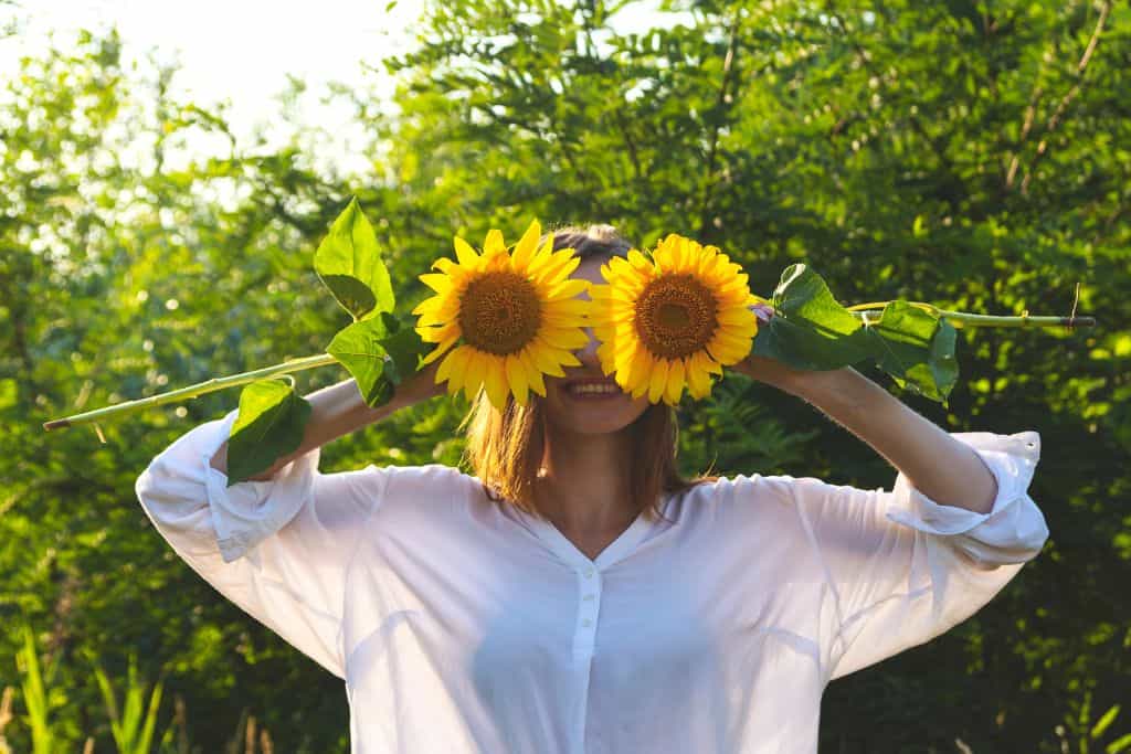 A smiling woman with two sunflowers covering her eyes.