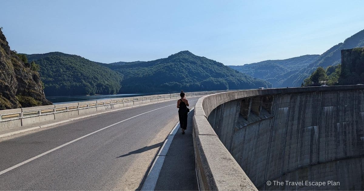 a lady walking along the Transfagarasan Highway on top of Vidraru Dam in Romania, with mountains in the distance